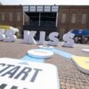 Kiss Kiss Way 2025: Villaggio con palco, stand brand e pubblico festante durante una tappa estiva del tour