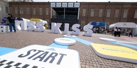 Kiss Kiss Way 2025: Villaggio con palco, stand brand e pubblico festante durante una tappa estiva del tour