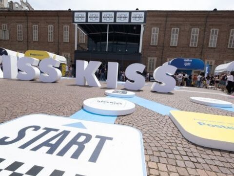 Kiss Kiss Way 2025: Villaggio con palco, stand brand e pubblico festante durante una tappa estiva del tour