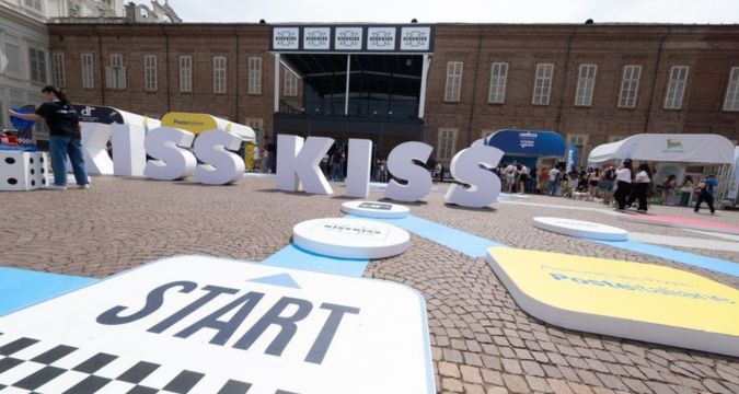 Kiss Kiss Way 2025: Villaggio con palco, stand brand e pubblico festante durante una tappa estiva del tour